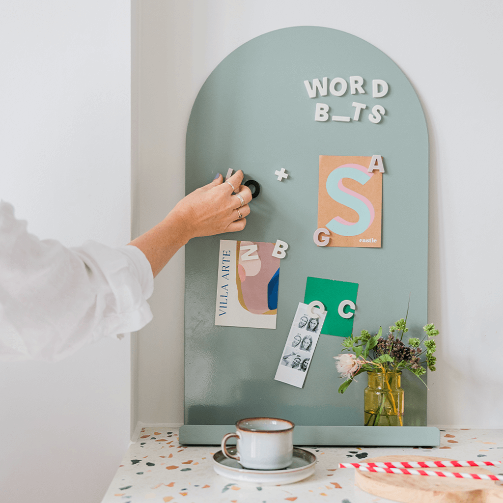 Sage green magnet board in modern kitchen on the countertop. Alphabet magnet letters on magnetic board with woman's hand placing a letter to hold up a postcard